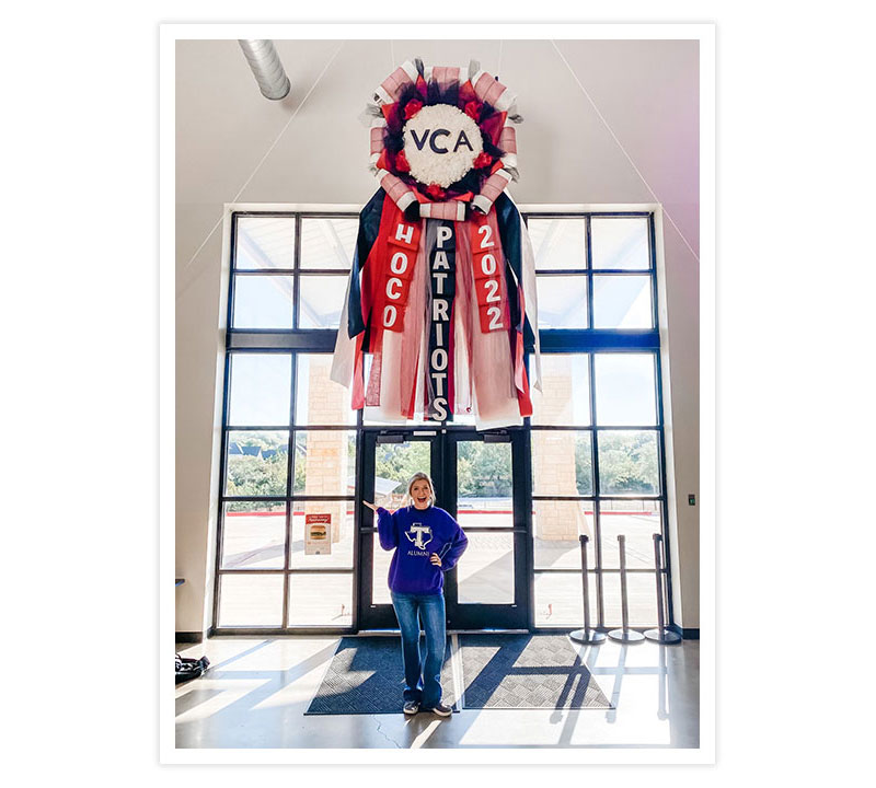 A woman with blonde hair holding her hand in the air wearing a purple Tarleton State University sweatshirt, posing with a giant Homecoming mum hanging on a wall.